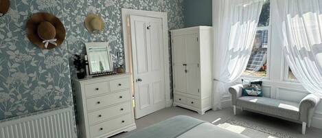 A bedroom with a large bed, a dresser, a wardrobe, a vanity with a mirror, and a window with sheer curtains.