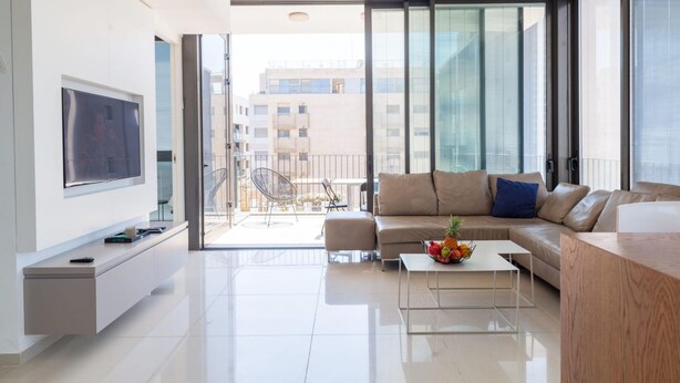 Ein modernes Wohnzimmer mit einem großen Flachbildfernseher, einem beigefarbenen Sofa, einem Couchtisch mit Obst und einem Balkon mit Blick auf Gebäude.