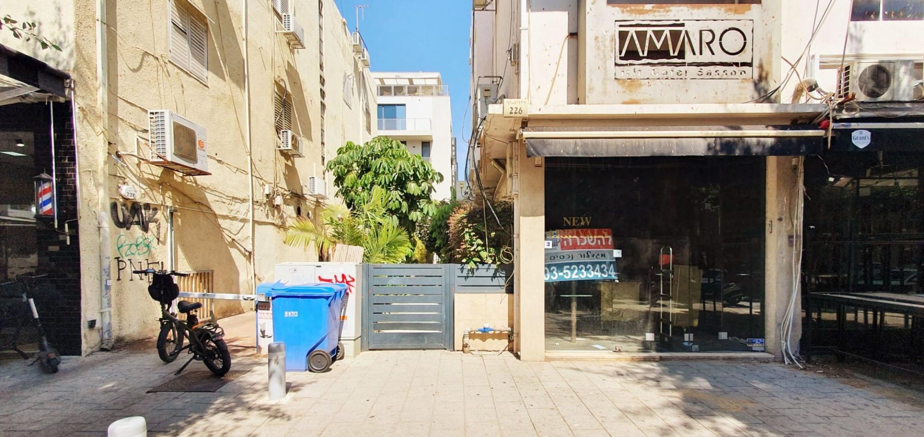 Una calle estrecha con una tienda cerrada llamada "AMARO" y un bote de basura azul al frente.