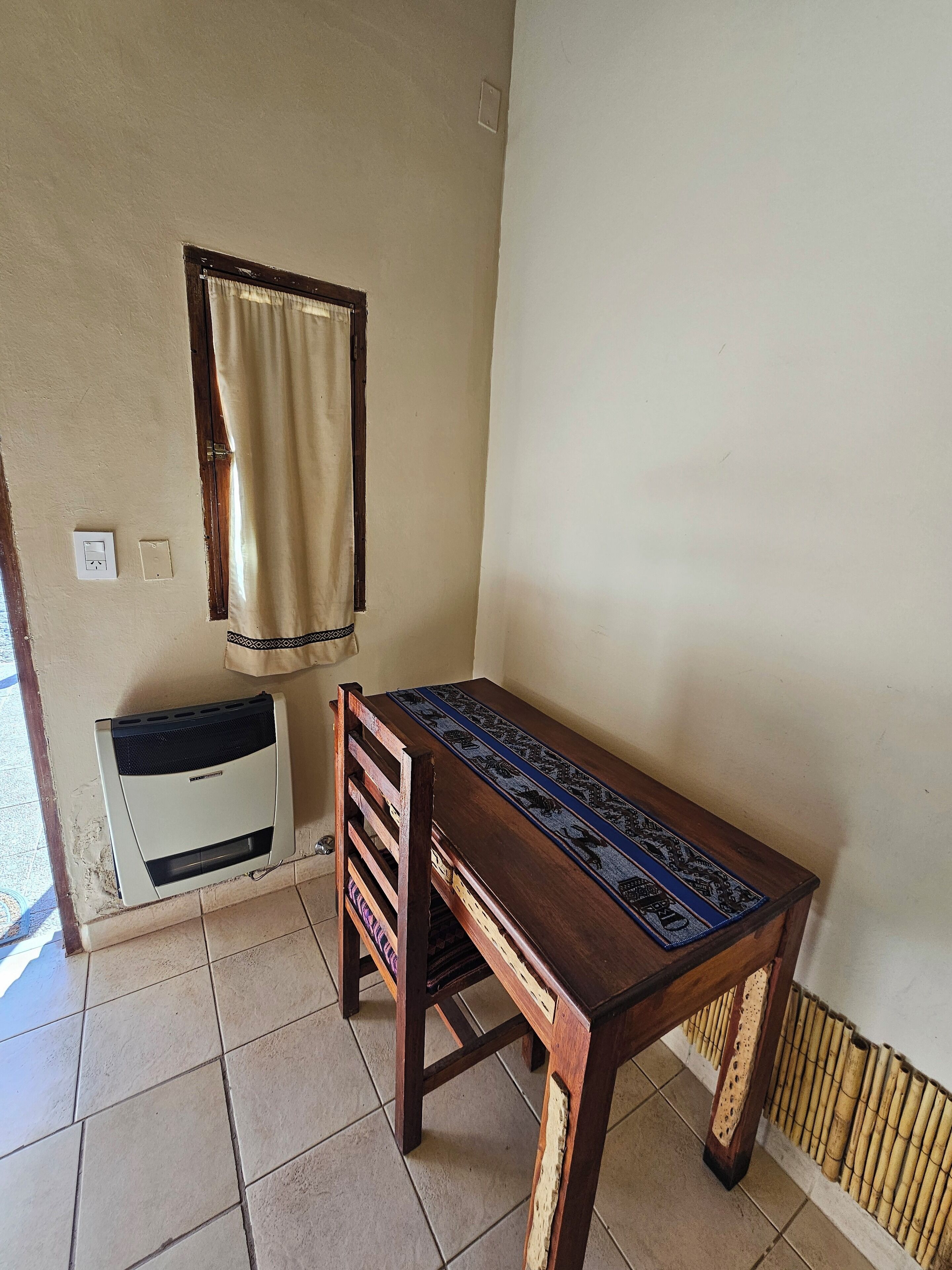 A small room with a wooden table and a chair, a window with a curtain, and a wall-mounted heater.