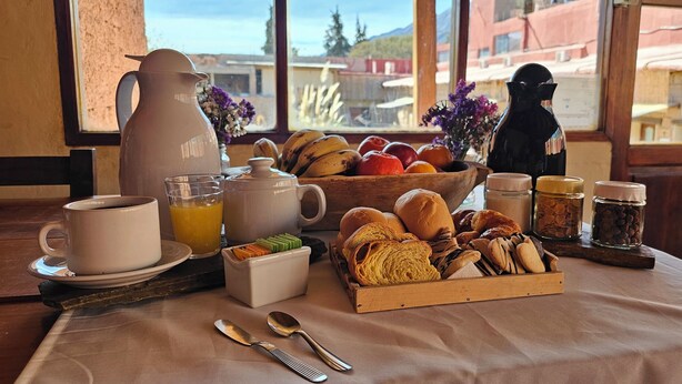 Une table dressée pour le petit-déjeuner, avec une cafetière, une théière, un panier de fruits, des viennoiseries et des condiments, et offrant une vue sur les bâtiments environnants.