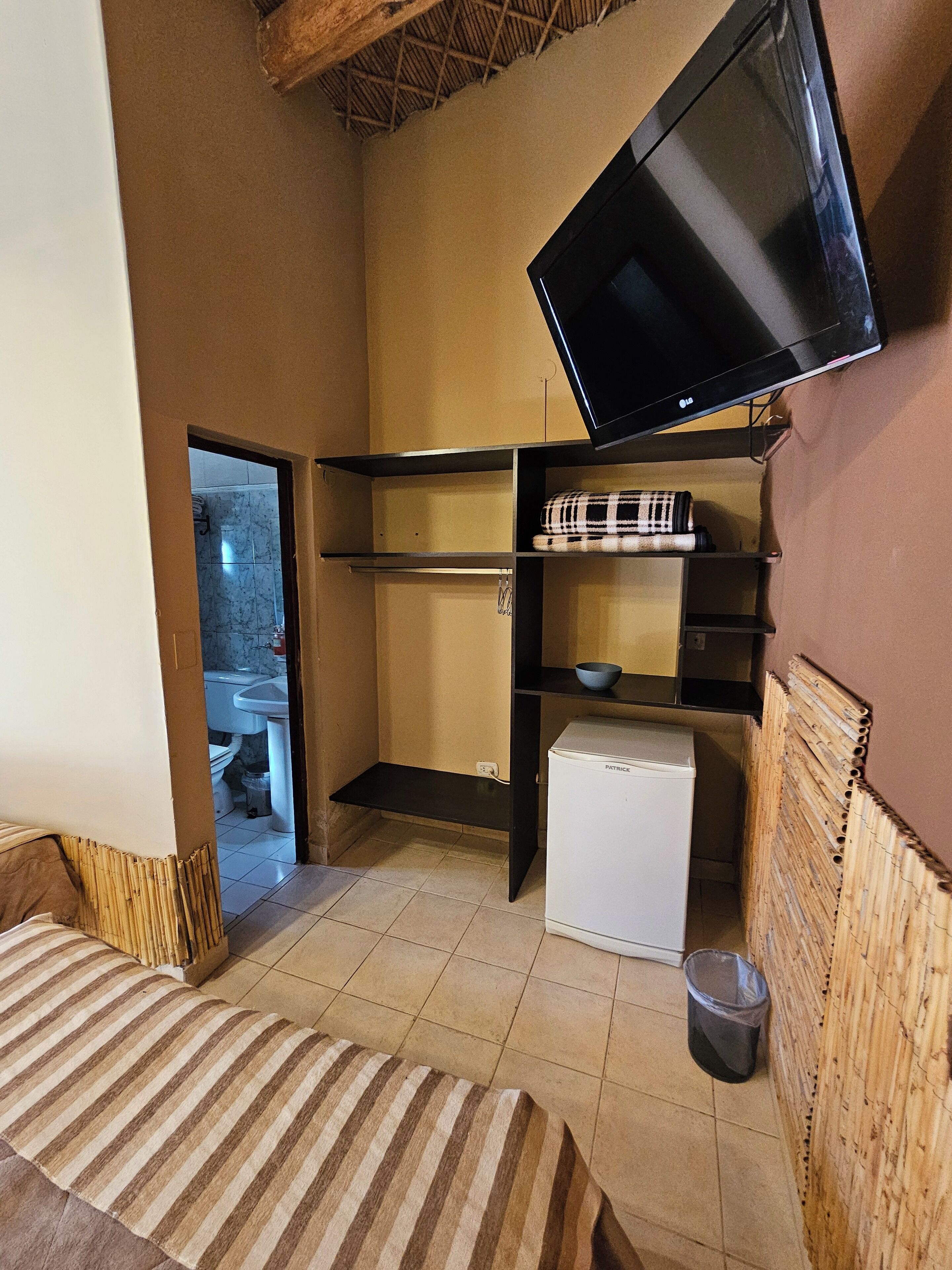 A compact room with a bed, a television mounted on the wall, a small refrigerator, and a shelf with a bowl.