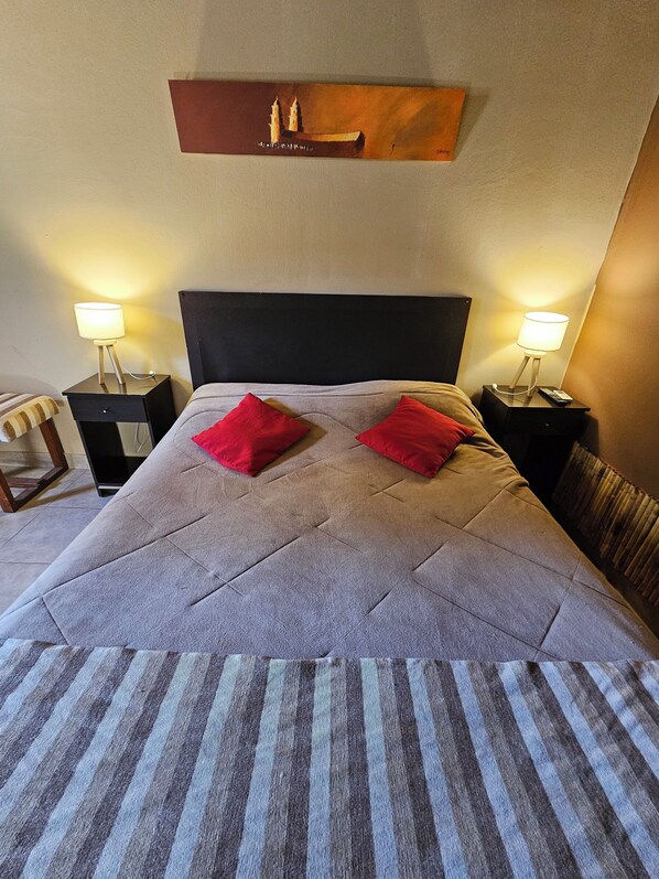 A neatly made bed with a striped blanket, two red pillows, and a headboard. There are two bedside tables with lamps and a small painting on the wall above the bed.