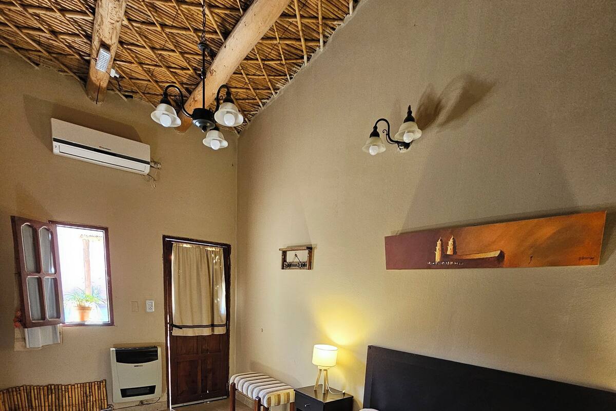 Une chambre à coucher comprenant un lit, un banc, une table de chevet avec une lampe, une étagère fixée au mur et une fenêtre avec des rideaux.