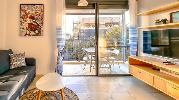 A modern living room with a black leather sofa, a white chair, a flat-screen TV, and a balcony with outdoor furniture. - Dizengoff Muguet in Tel Aviv-Yafo (Tel Aviv-Yafo)