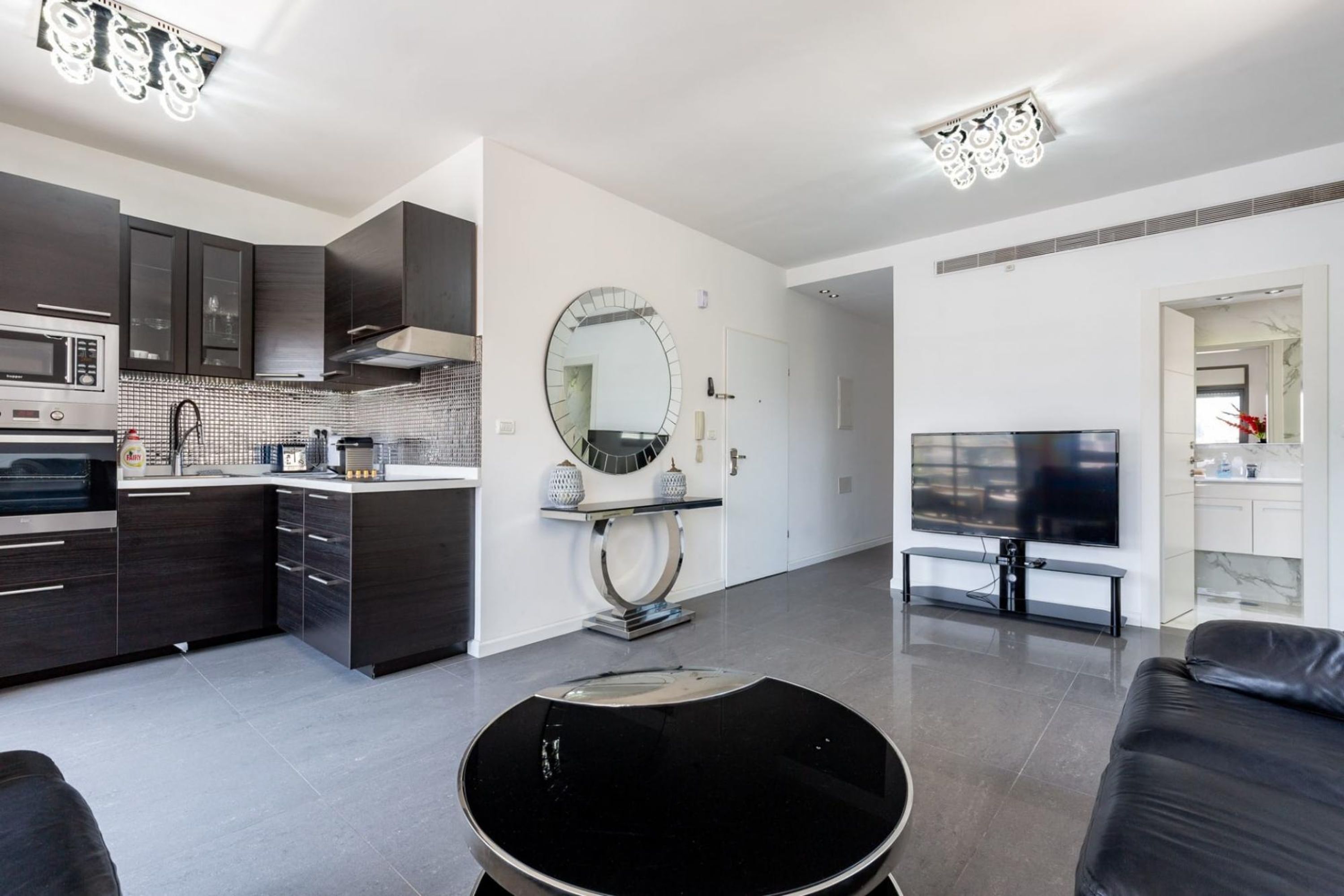 A modern kitchen with dark cabinetry, a microwave, and a built-in oven. There is a round mirror on the wall and a flat-screen TV in the living area.