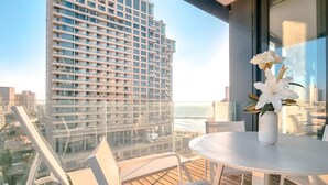Een balkon met een witte tafel en stoelen, een vaas met witte bloemen, en uitzicht op een hoog gebouw en de oceaan.
