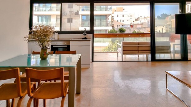 Eine moderne Küche mit einem Glastisch und Stühlen, einer Vase mit Trockenblumen und Blick auf die Stadt durch große Fenster.