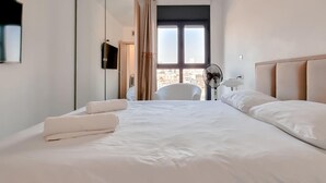 Ein Hotelzimmer mit einem großen Bett, einem Sessel, einem Ventilator und Blick auf die Stadt durch ein Fenster.