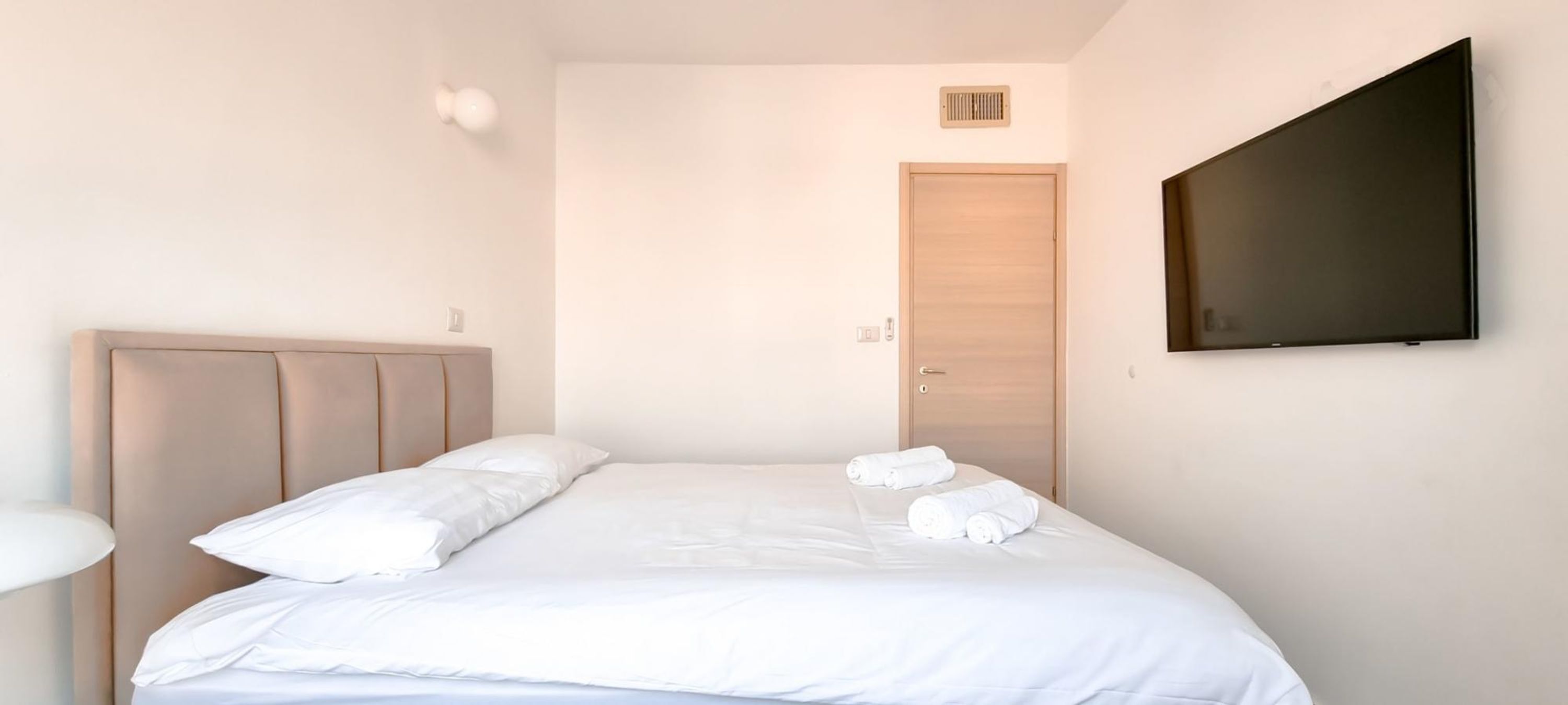 Ein ordentlich bezogenes Bett mit weißen Leinen und Handtüchern, ein Holz-Kopfteil, ein an der Wand befestigtes Fernsehgerät und eine Tür im Hintergrund.