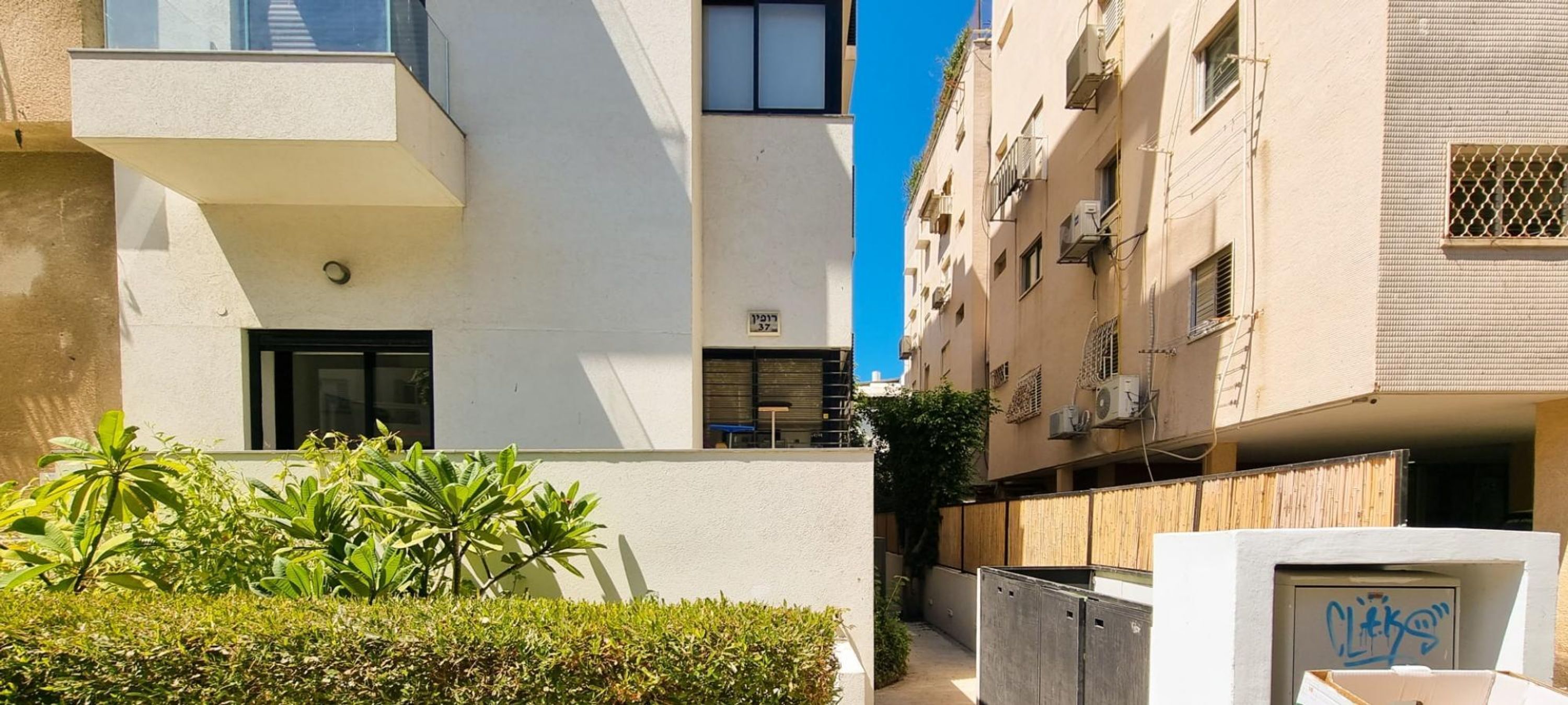 A modern apartment building with balconies, a small garden area, and a visible door.