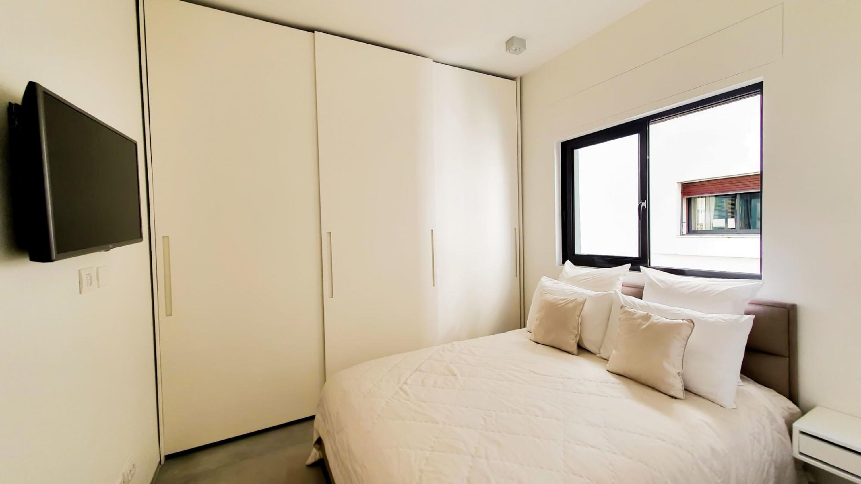 A bedroom with a bed, a TV mounted on the wall, and a window with a view of the outside.