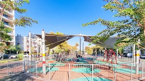 Une aire de jeux avec des balançoires et un espace ombragé, entourée d’une clôture et d’arbres.