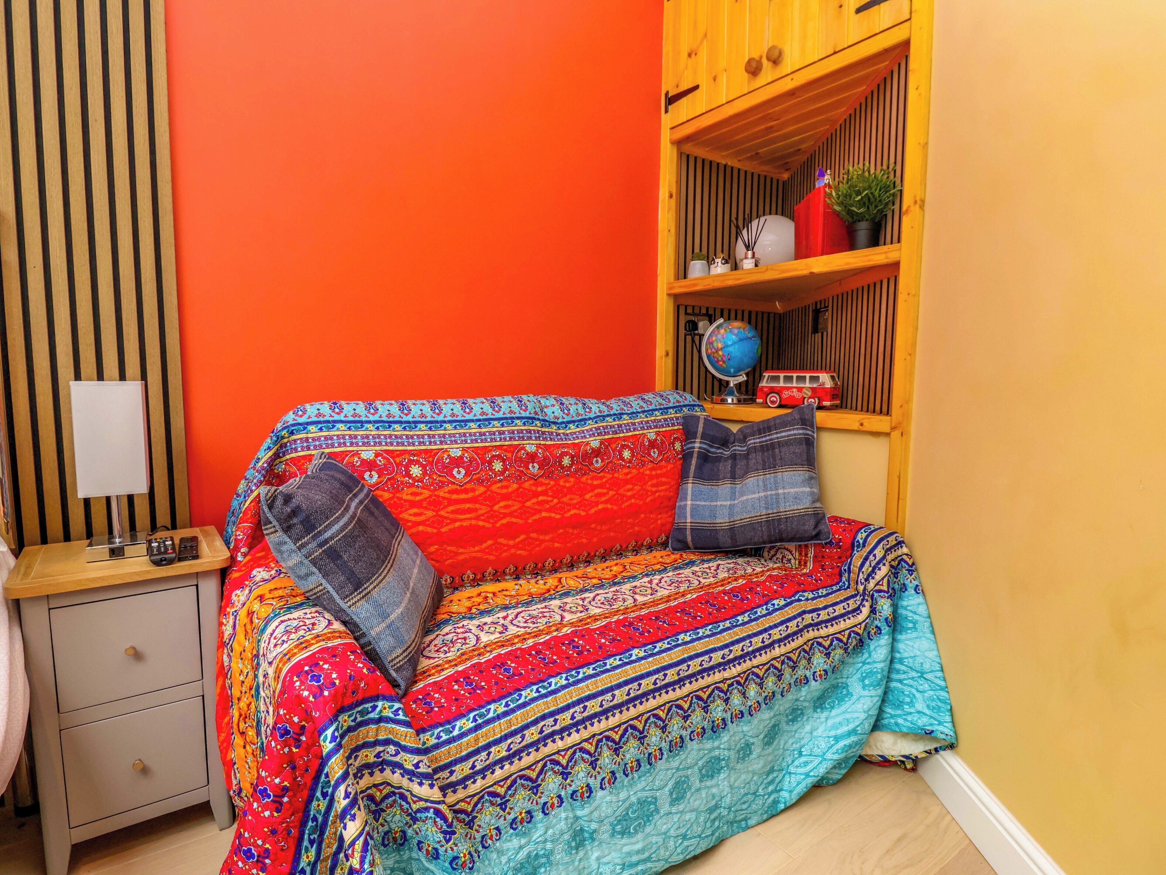 A room with a colorful patterned sofa, a small wooden shelf with decorative items, a lamp, and a bedside table.