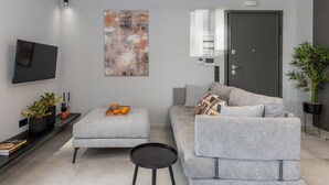 A modern living room with a grey sofa, a small ottoman, a black coffee table, a flat-screen TV, a large abstract painting, a door with a security panel, and a potted plant. - Superior Suite with sea views to Argostoli bay (Argostoli)