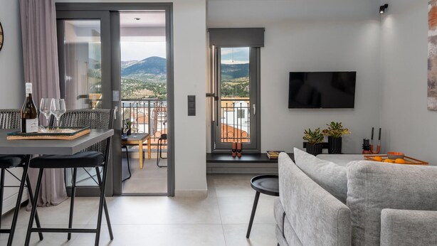 A modern living room with a sofa, a dining area with a table and chairs, a balcony with a view of the mountains, and a flat-screen TV mounted on the wall. - Superior Suite with sea views to Argostoli bay (Argostoli)