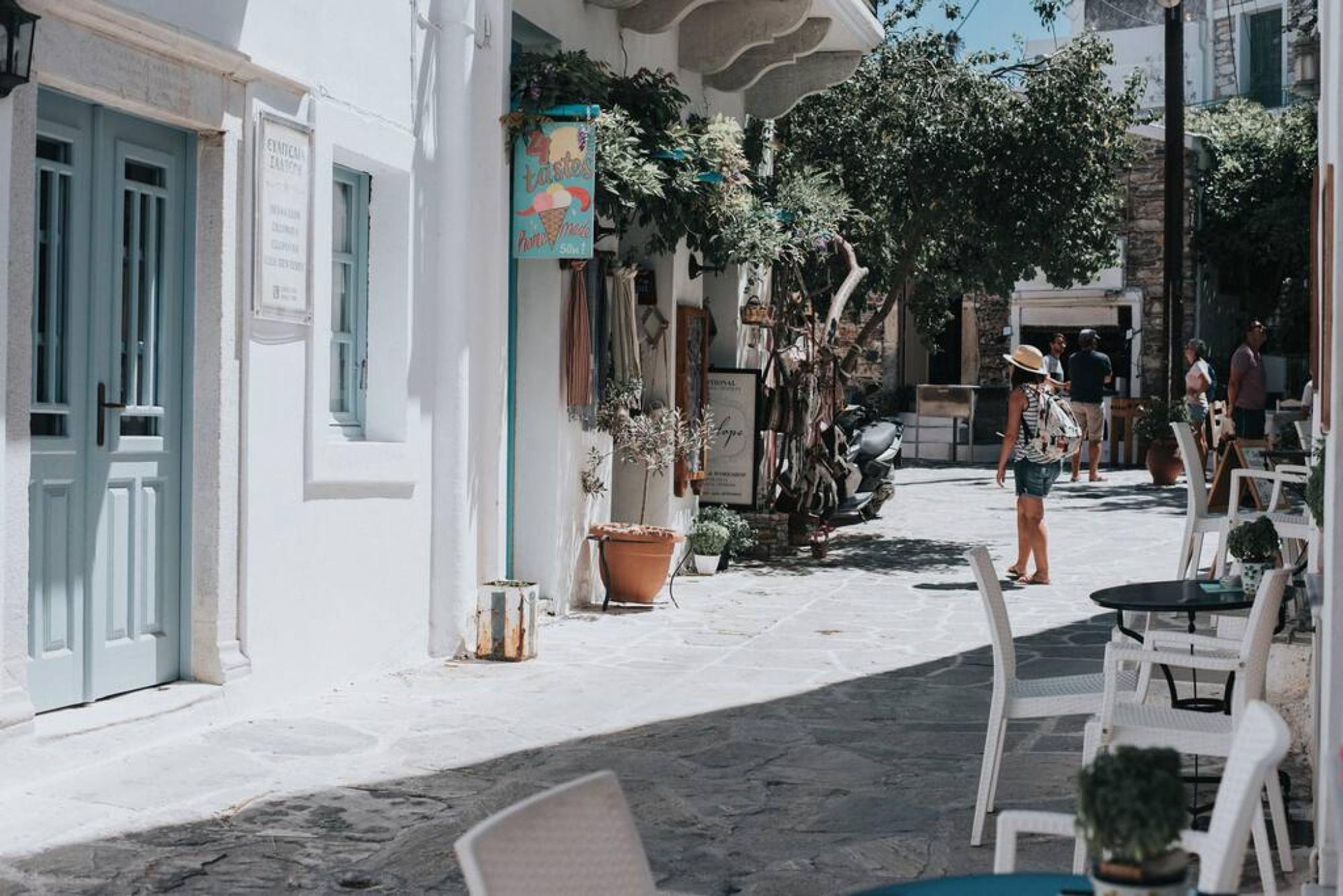 Una calle estrecha con edificios blancos, mesas al aire libre y plantas en macetas.