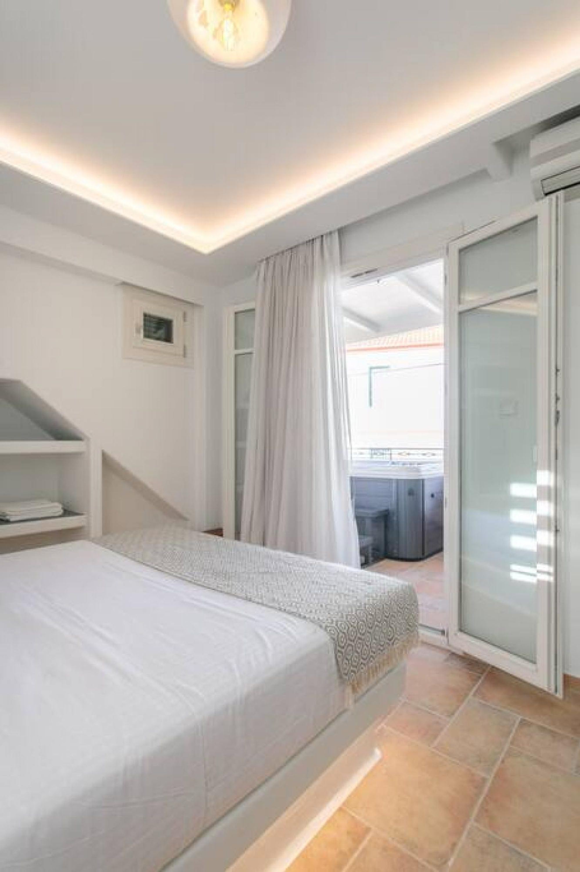 A modern bedroom with a large bed, a built-in bench, a small bedside table, and a sliding glass door leading to a balcony.