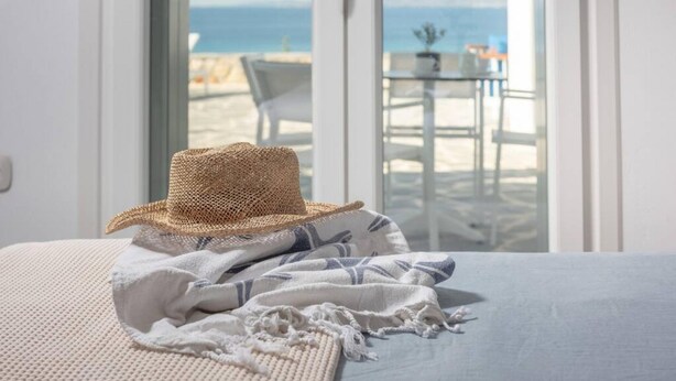 A bed with a straw hat and a white towel, a view of a beach and ocean through a glass door, and a small table with chairs outside. - Villa Agali Naxos Glyfada Kastraki (Naxos)
