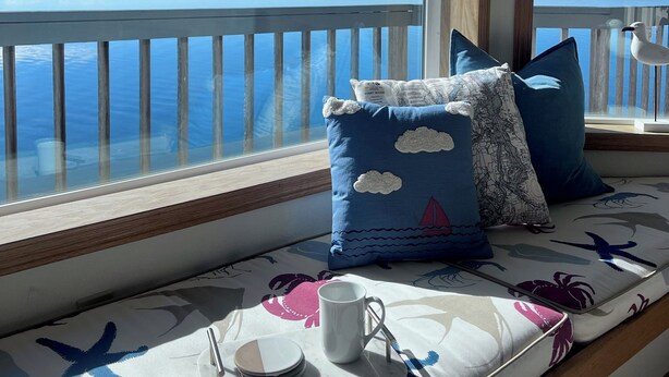 A balcony with a view of a calm body of water, a wooden railing, and a seating area with cushions and a tray with a mug and plates. - Fox Island Beachfront Retreat - Kayaks, Canoe, Paddle Board & Epic Sunsets (Fox Island)