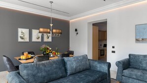 Una sala de estar moderna con un sofá gris, una mesa de comedor con frutas y bebidas, y una cocina al fondo.