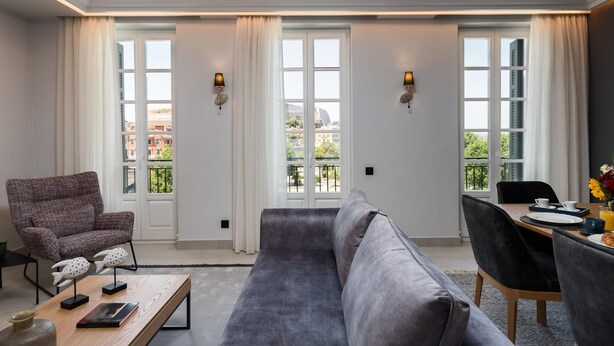 Une chambre d’hôtel moderne avec un canapé gris, une table à manger avec des chaises et une vue sur la ville grâce à de grandes fenêtres.