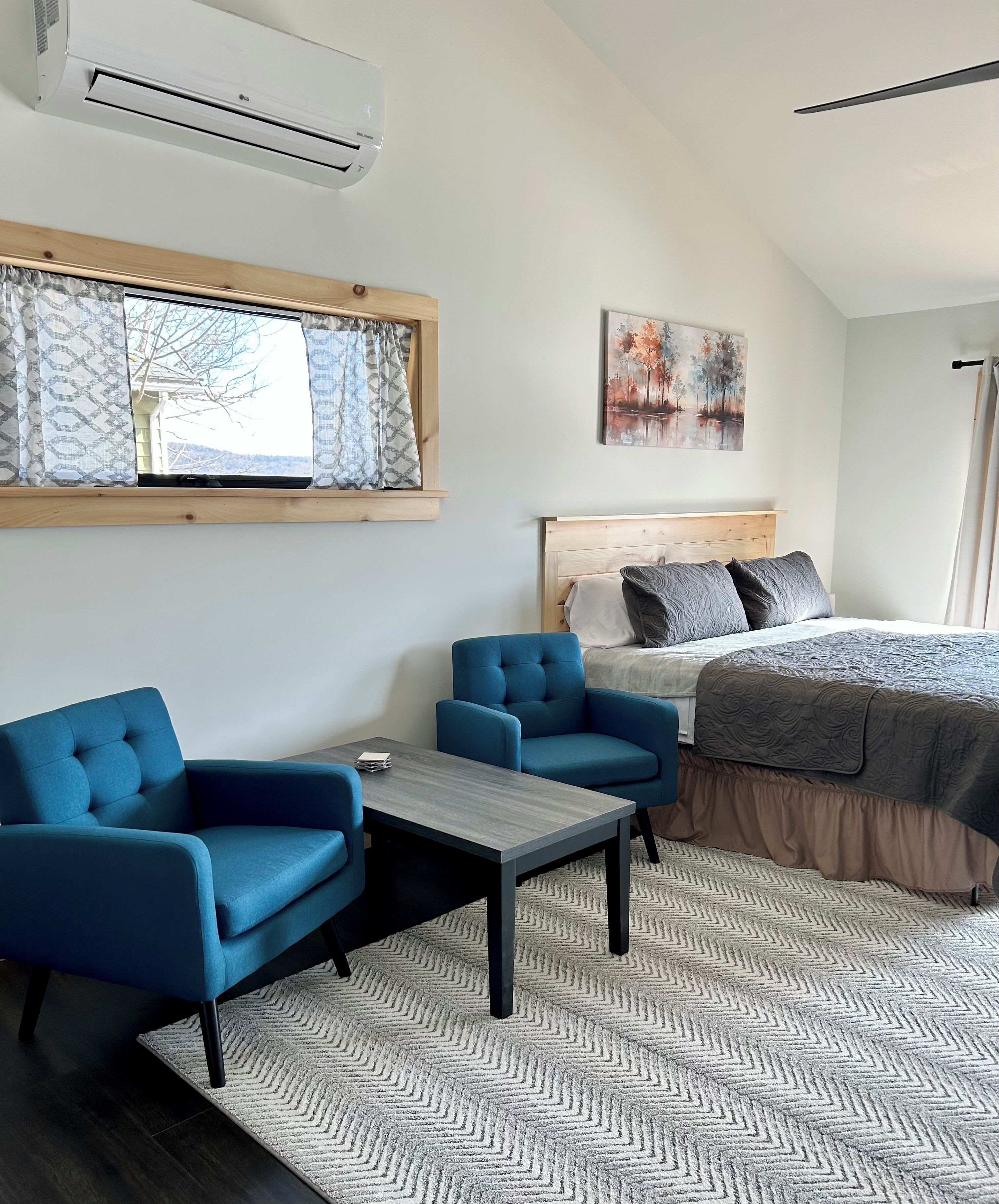 Une chambre à coucher avec un lit, deux fauteuils bleus, une petite table, une fenêtre avec des rideaux à motifs, un climatiseur et un tableau accroché au mur.
