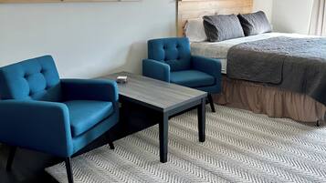Une chambre à coucher avec un lit, deux fauteuils bleus, une petite table, une fenêtre avec des rideaux à motifs, un climatiseur et un tableau accroché au mur.