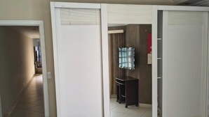 Ein Hotelzimmer mit Bett, Kleiderschrank, einem kleinen Tisch und einem Spiegel. Ein Flur führt zu einem weiteren Zimmer.