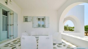 A room with a white interior, a dining area with a glass table and white chairs, and a view of the outdoors through an arched window. - Angel Villa in Paros (Lagkeri)