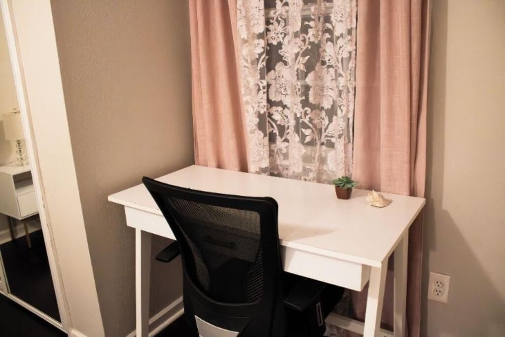 A small, white desk with a black mesh chair, a potted plant, and a window with floral curtains.