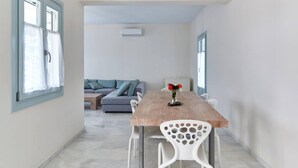A dining area with a wooden table, white chairs, and a vase of flowers. There is a gray sofa and a small table in the background. - Aegean Villa in Paros (Lagkeri)