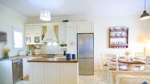 Cozinha moderna com armários brancos, uma ilha central, eletrodomésticos de aço inoxidável e uma área de jantar com mesa e cadeiras de madeira.