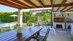 Um pátio externo com mesa e cadeiras de madeira, churrasqueira de tijolos e uma piscina ao fundo.