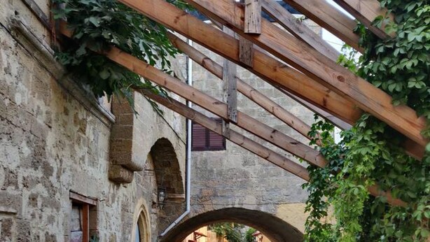 A narrow cobblestone alley with a wooden archway, stone buildings, and potted plants. - Megiston House in Old Town Rhodes (Rodos)