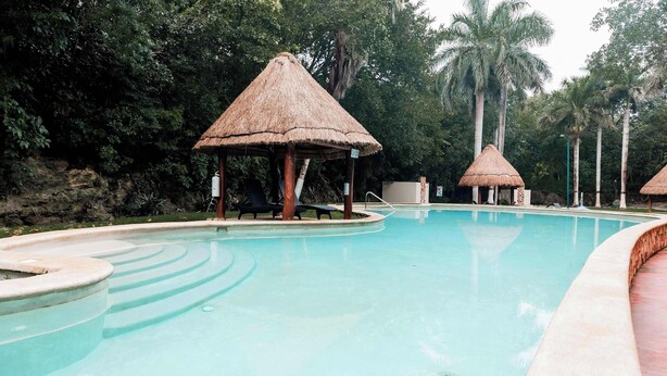 Ein Swimmingpool mit strohgedeckten Gebäuden, umgeben von üppiger Vegetation.