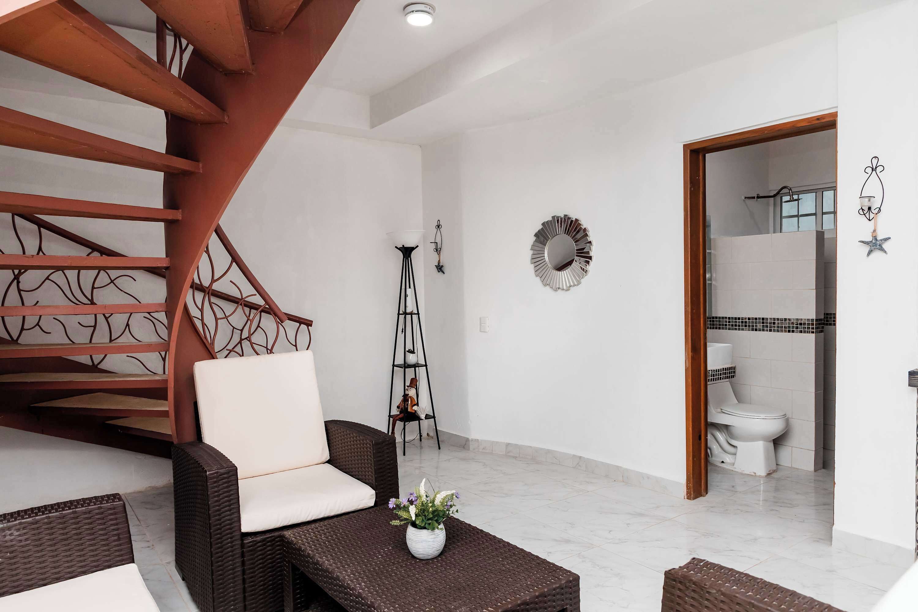 Un interior moderno con una escalera, muebles de mimbre, una pequeña mesa con una planta y un baño visible a través de una puerta abierta.