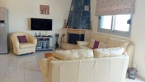 Una sala de estar con un sofá beige, un sillón pequeño, un televisor de pantalla plana, una chimenea y una ventana con cortinas estampadas.
