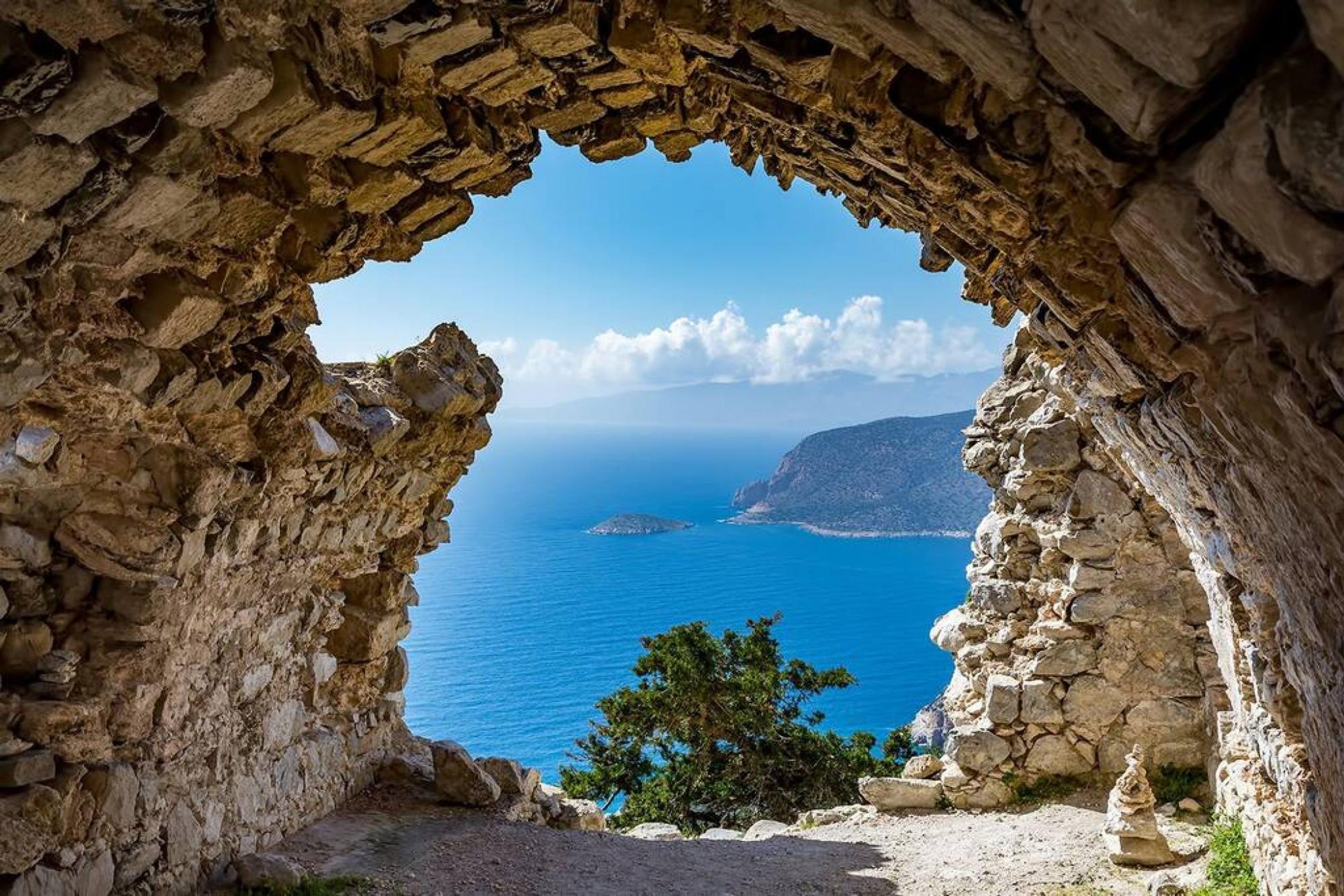 Ein steinerner Bogen führt zu einer Küstenansicht mit klarem, blauem Meer und weiter Landpartie.