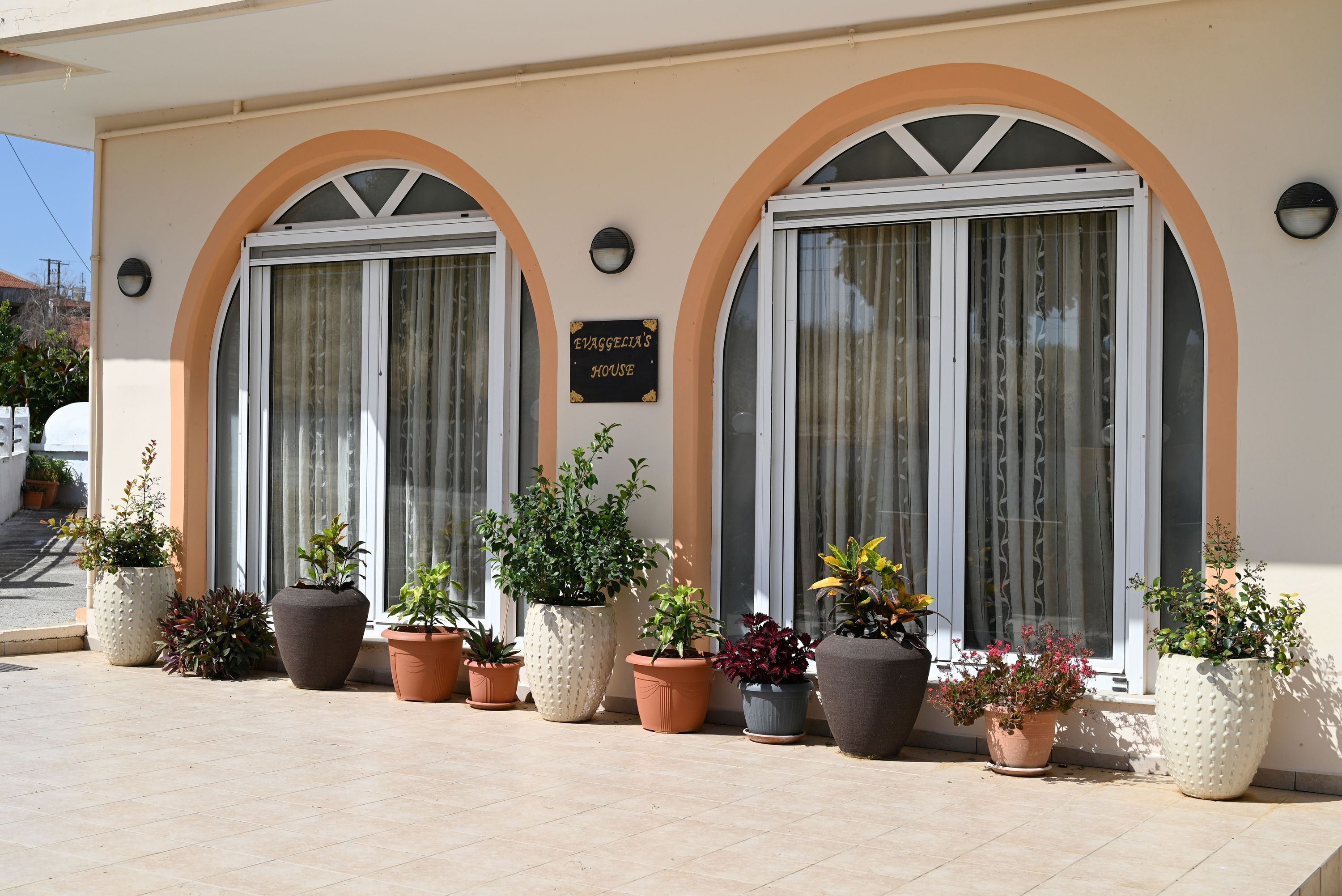 A building with two large arched windows, a sign reading "EVANGELICAL HOUSE," and several potted plants in front.