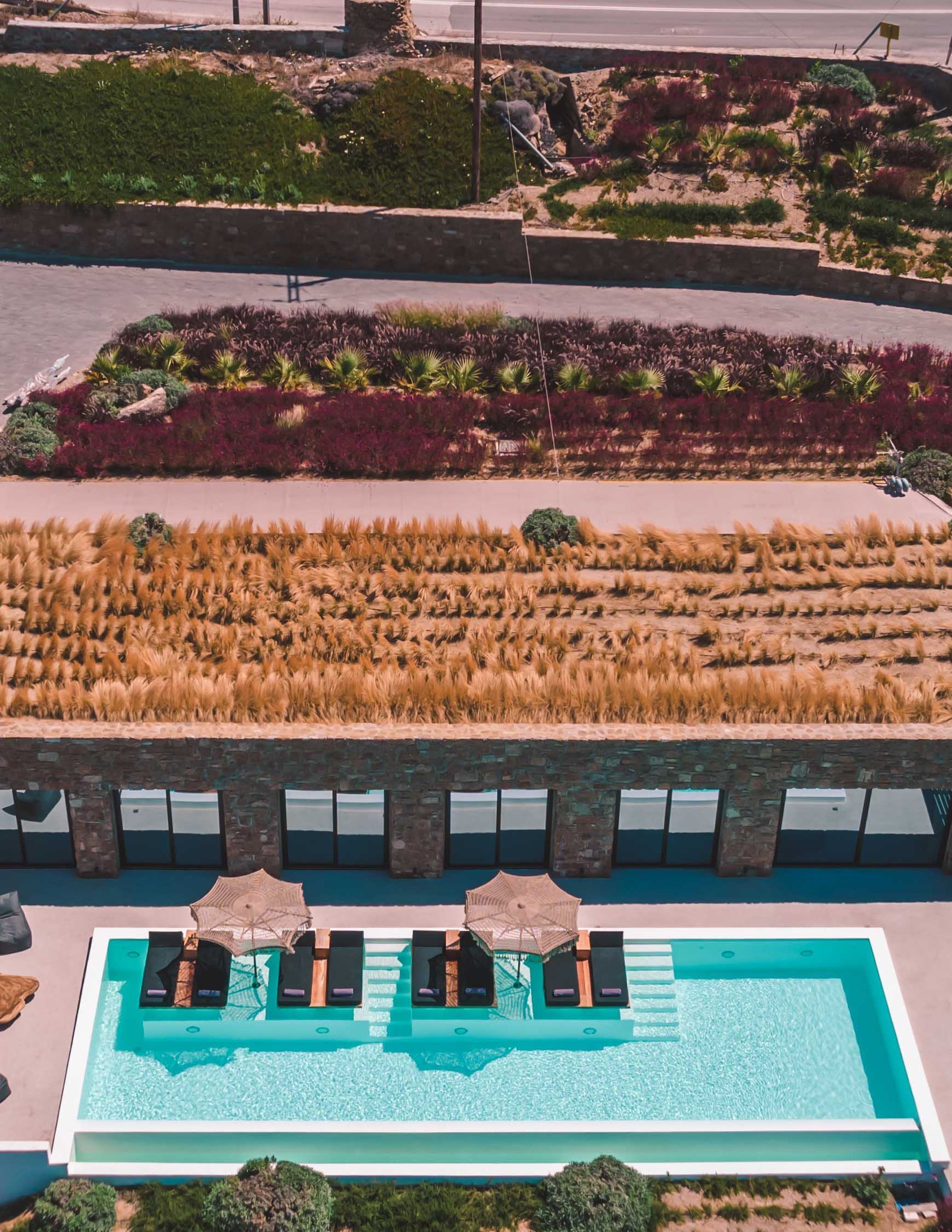 An aerial view of a modern hotel with a rectangular pool, outdoor seating with umbrellas, and landscaped gardens.
