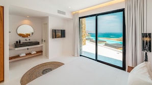 A bedroom with a large bed, a round mirror, a modern bathroom with a sink and towel racks, and a view of a pool and ocean through floor-to-ceiling windows. - Sea la vie villa Private pool (MYKONOS)