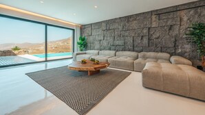 A modern living room with a large window, a stone wall, a beige sofa, and a round coffee table. - Sea la vie villa Private pool (MYKONOS)