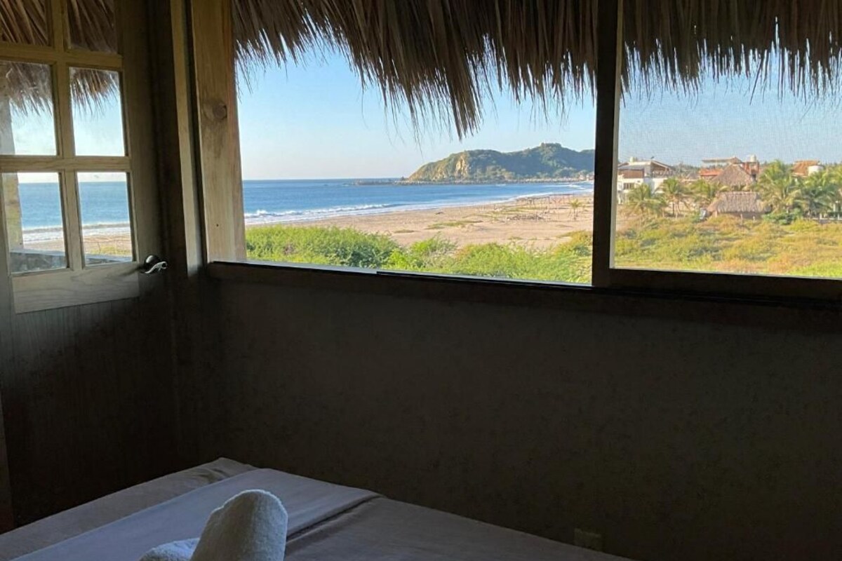 Une chambre avec un lit, une fenêtre donnant sur la plage et les montagnes, et un toit de chaume.