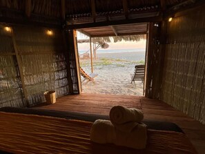 Une pièce avec un plancher en bois, un lit sur lequel est posée une serviette roulée, une fenêtre donnant sur une plage et une structure avec un toit de chaume.