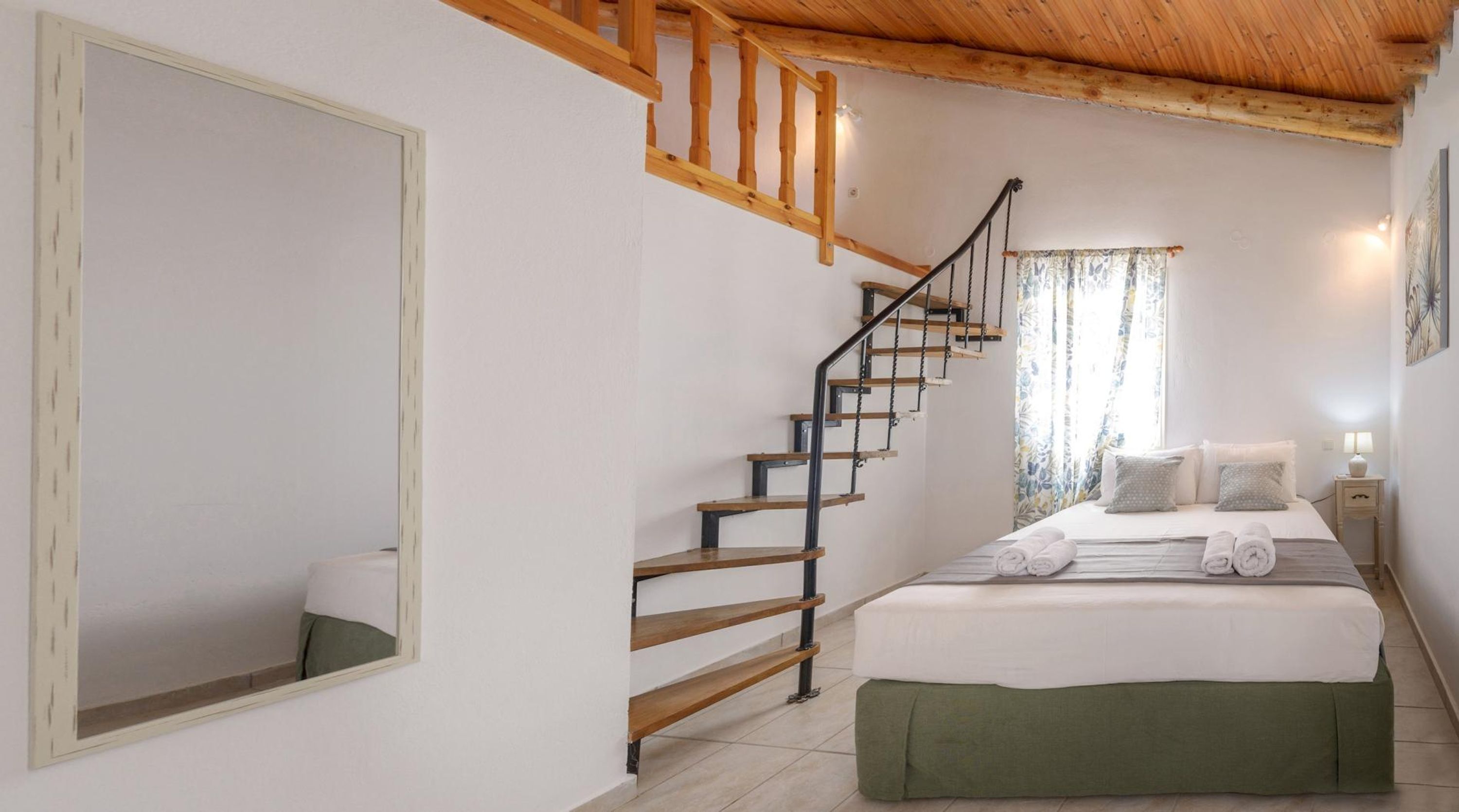 Une chambre avec un escalier en bois, un grand miroir, un lit avec des serviettes pliées et une table de chevet avec une lampe.