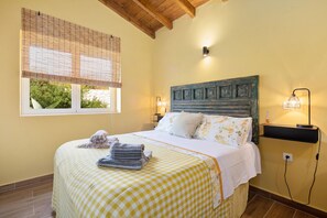 A bedroom with a checkered bedspread, a wooden headboard, two bedside lamps, and a window with blinds. - Bibi - Danilia Cottages (Gaios)
