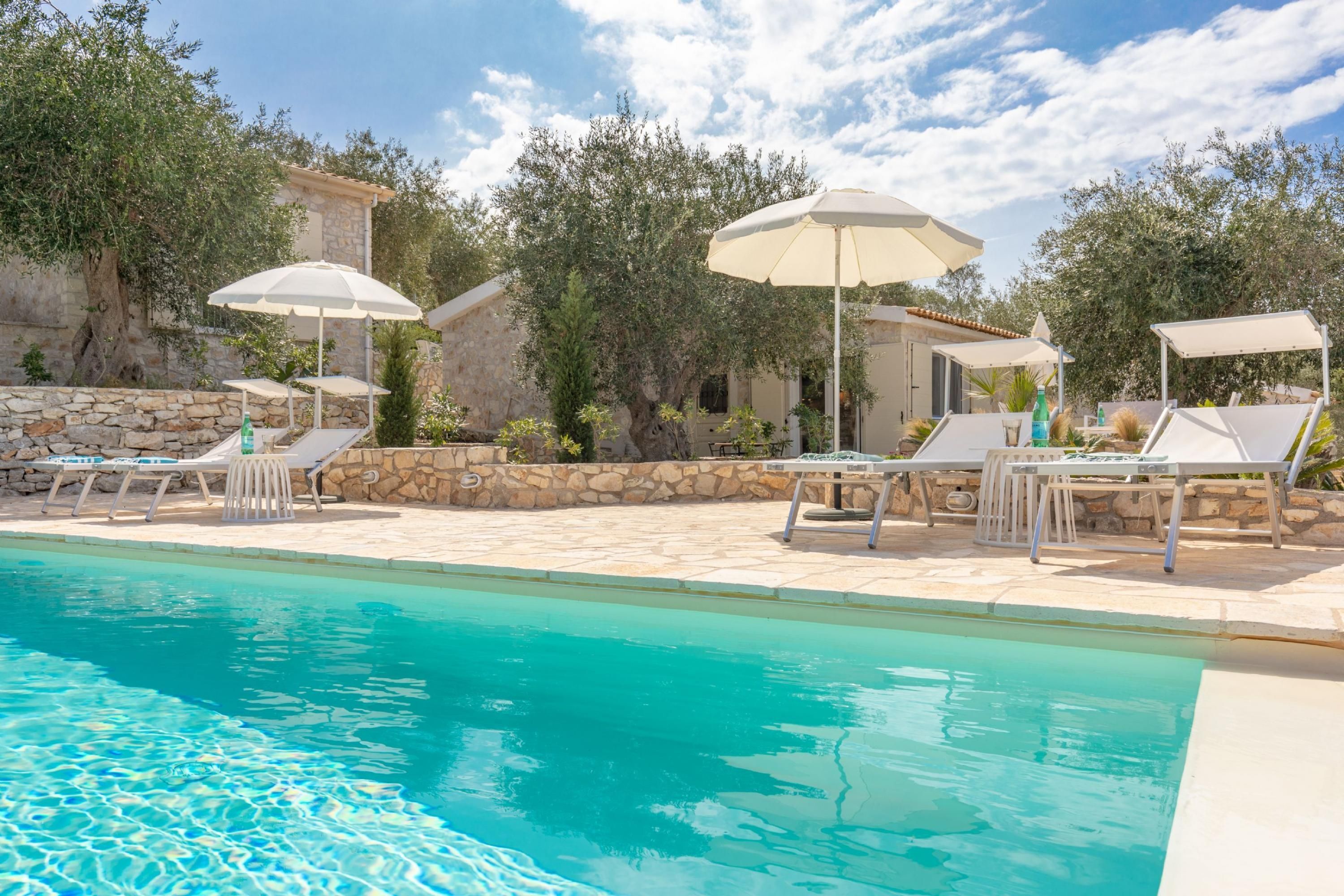 Une piscine entourée de murs de pierre et d’arbres, avec des chaises longues et des parasols.