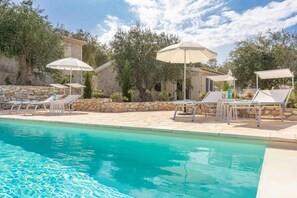 Una piscina con sillones de descanso y sombrillas, rodeada de muros de piedra y árboles.