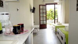 A kitchen with a coffee maker, mugs, and glasses on the counter. There is a dining area with a table and chairs, and a bedroom with a bed and a window.